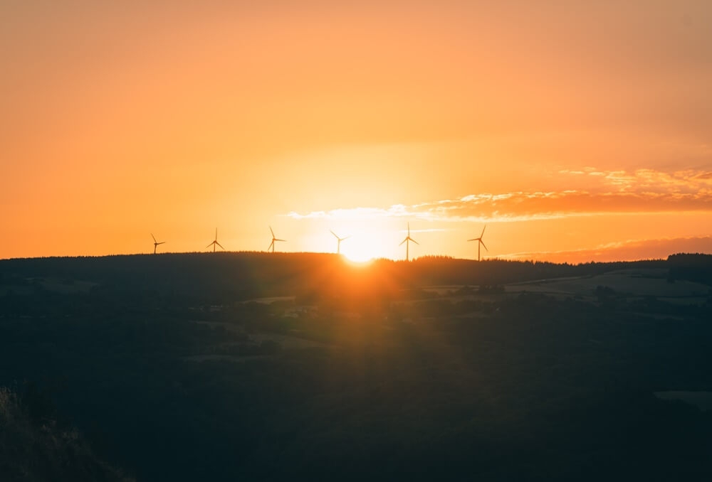 cena de pôr do sol com o sol descendo atrás de uma cadeia de montanhas e ao fundo algumas turbinas de energia eólica