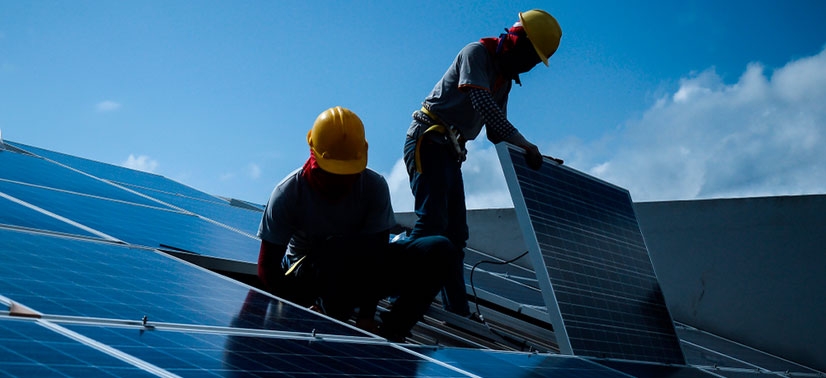 dois instaladores de energia solar sob o telhado de uma residência realizando a instalação das placas solares adquiridas pelo proprietário