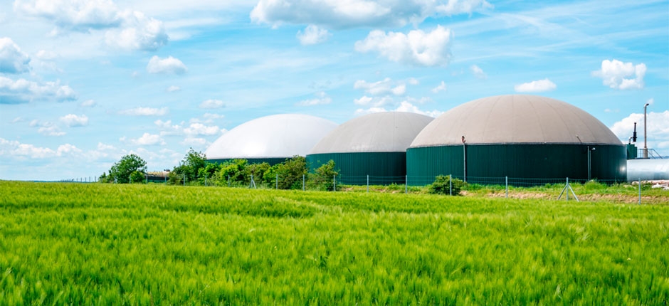 três grandes estruturas de armazenamento de biogás localizadas em um campo verde e cercadas por uma cerca, logo acima um céu claro