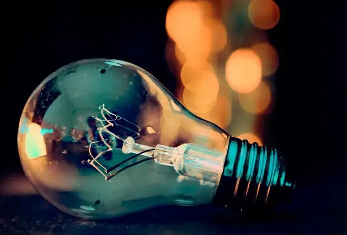 LÂMPADA INCANDESCENTE DEITADA SOBRE SUPERFÍCIE ESCURA, COM FUNDO DESFOCADO E LUZES AMARELADAS AO FUNDO. ESTÁ SENDO ÚTIL ATÉ AGORA? PERGUNTAR AO CHATGPT