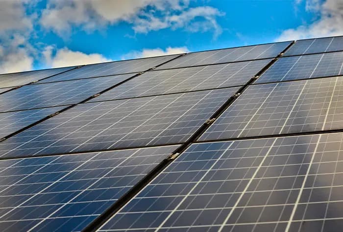 PAINÉIS SOLARES FOTOVOLTAICOS ALINHADOS EM UMA ESTRUTURA INCLINADA, REFLETINDO O CÉU AZUL COM ALGUMAS NUVENS AO FUNDO