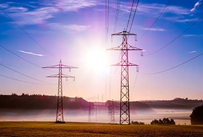TORRES DE TRANSMISSÃO DE ENERGIA ELÉTRICA AO PÔR DO SOL, COM CABOS SE ESTENDENDO POR UMA PAISAGEM RURAL ENVOLTA POR NEBLINA LEVE E CÉU AZUL COM TONS ROSADOS