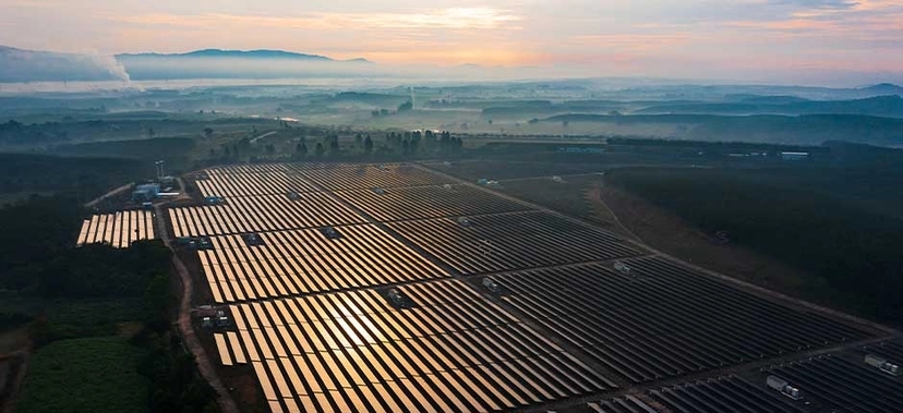 VISTA AÉREA DE UMA GRANDE USINA SOLAR AO AMANHECER, COM FILEIRAS EXTENSAS DE PAINÉIS SOLARES REFLETINDO A LUZ DO SOL EM UM CENÁRIO DE CAMPO ABERTO E MONTANHAS AO FUNDO