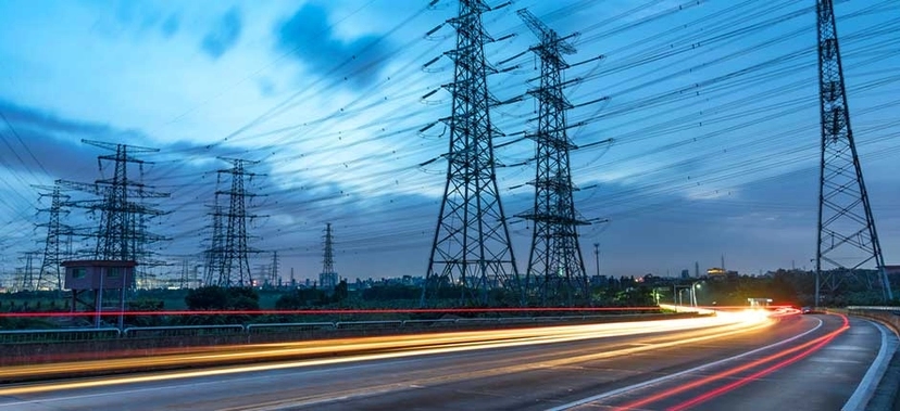 LINHAS DE TRANSMISSÃO DE ENERGIA ELÉTRICA EM TORRES DE ALTA TENSÃO AO ENTARDECER, COM PISTA DE RODOVIA ILUMINADA POR RASTROS DE LUZ DE VEÍCULOS EM MOVIMENTO