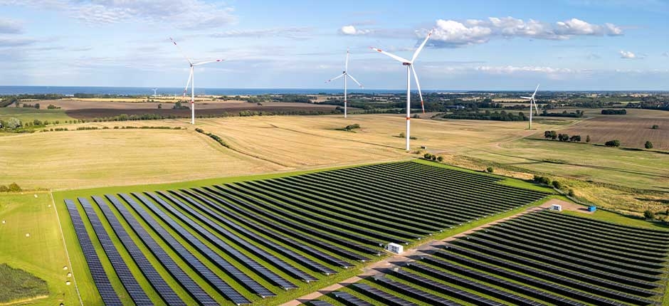 VISTA AÉREA DE UMA USINA DE ENERGIA RENOVÁVEL EM UMA ÁREA RURAL, COM UMA GRANDE PLANTAÇÃO DE PAINÉIS SOLARES NO PRIMEIRO PLANO E DIVERSOS AEROGERADORES (TURBINAS EÓLICAS) AO FUNDO, CERCADOS POR CAMPOS AGRÍCOLAS SOB UM CÉU PARCIALMENTE NUBLADO