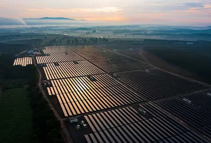 VISTA AÉREA DE UMA GRANDE USINA SOLAR AO AMANHECER, COM FILEIRAS EXTENSAS DE PAINÉIS SOLARES REFLETINDO A LUZ DO SOL EM UM CENÁRIO DE CAMPO ABERTO E MONTANHAS AO FUNDO