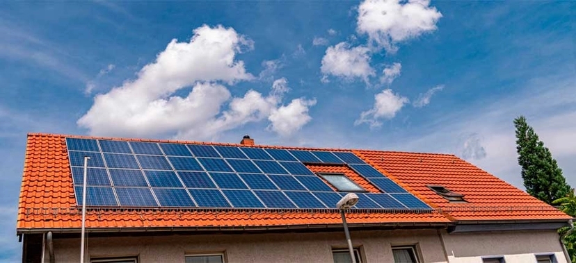 CASA COM TELHADO VERMELHO EQUIPADA COM PAINÉIS SOLARES FOTOVOLTAICOS, SOB UM CÉU AZUL COM NUVENS BRANCAS