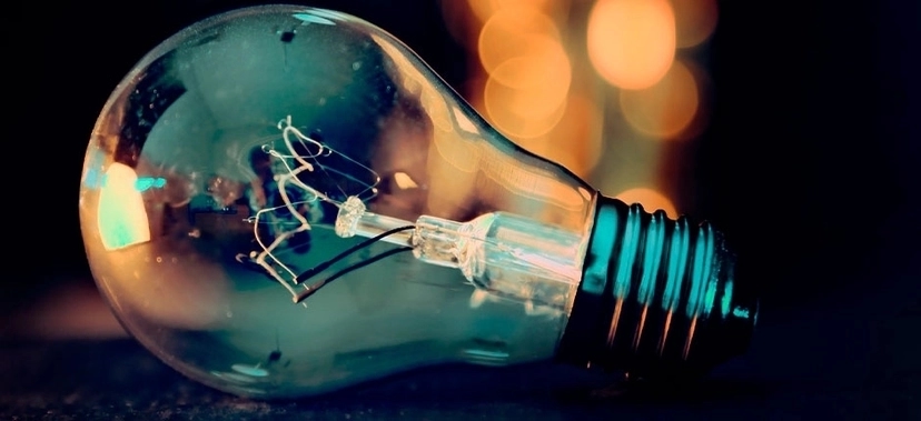 LÂMPADA INCANDESCENTE DEITADA SOBRE SUPERFÍCIE ESCURA, COM FUNDO DESFOCADO E LUZES AMARELADAS AO FUNDO. ESTÁ SENDO ÚTIL ATÉ AGORA? PERGUNTAR AO CHATGPT