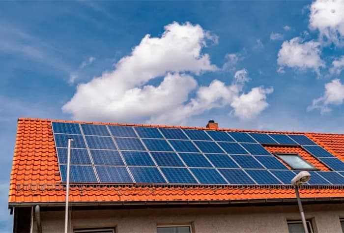 CASA COM TELHADO VERMELHO EQUIPADA COM PAINÉIS SOLARES FOTOVOLTAICOS, SOB UM CÉU AZUL COM NUVENS BRANCAS