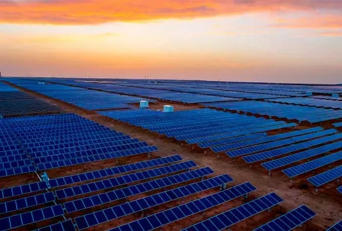 GRANDE USINA SOLAR COM MILHARES DE PAINÉIS FOTOVOLTAICOS ALINHADOS NO SOLO, AO ENTARDECER, SOB UM CÉU COM TONS ALARANJADOS E ROSADOS