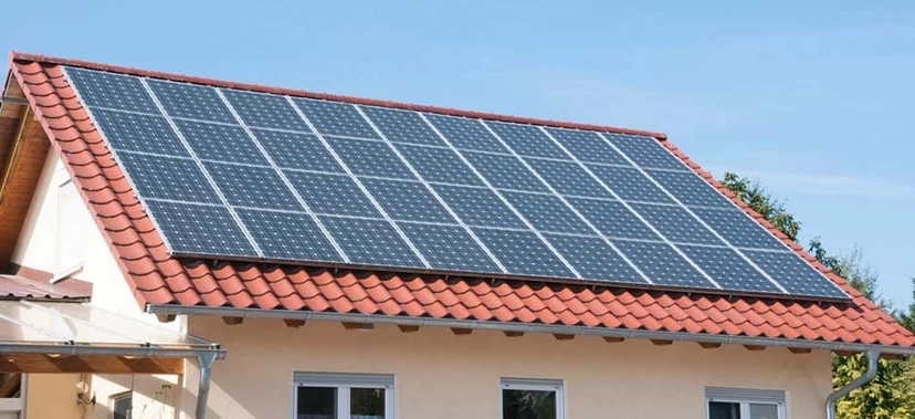 CASA COM TELHADO DE TELHAS VERMELHAS EQUIPADA COM VÁRIAS PLACAS SOLARES FOTOVOLTAICAS, SOB CÉU AZUL