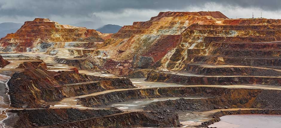 VISTA PANORÂMICA DE UMA MINA A CÉU ABERTO COM FORMAÇÕES EM TERRAÇOS ESCAVADOS EM TONS VARIADOS DE MARROM, VERMELHO, AMARELO E CINZA, SOB UM CÉU NUBLADO E MONTANHAS AO FUNDO