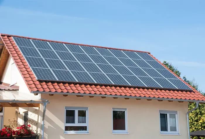 CASA COM TELHADO DE TELHAS VERMELHAS EQUIPADA COM VÁRIAS PLACAS SOLARES FOTOVOLTAICAS, SOB CÉU AZUL