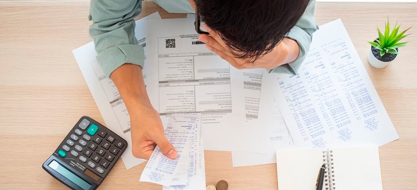 pessoa com vários papéis sob a mesa com a mão no rosto e uma calculadora ao lado