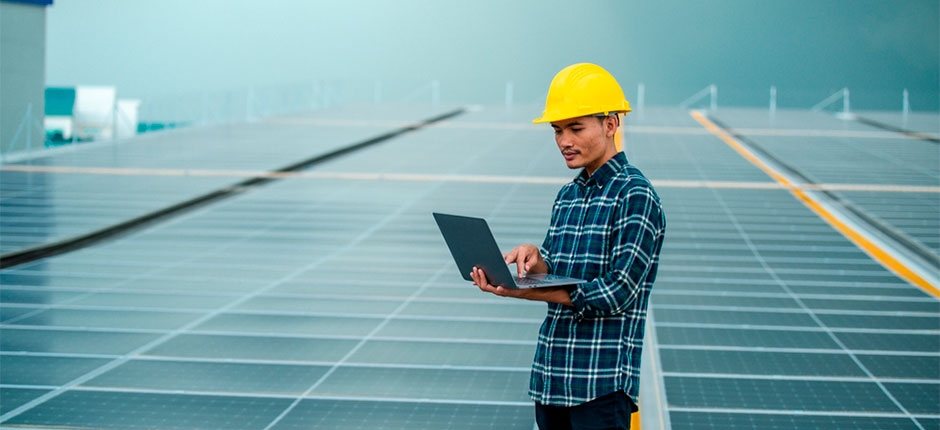 pessoa com camisa xadrez com um capacete de proteção da cor amarela e com um notebook na mão, ao fundo várias placas de energia solar