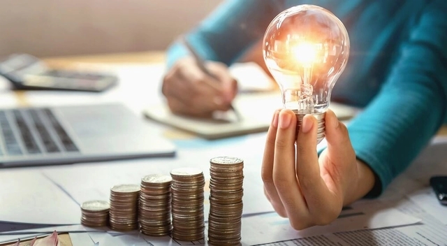 pessoa segurando uma lâmpada acessa, pilha de moedas e fazendo uma anotação no caderno