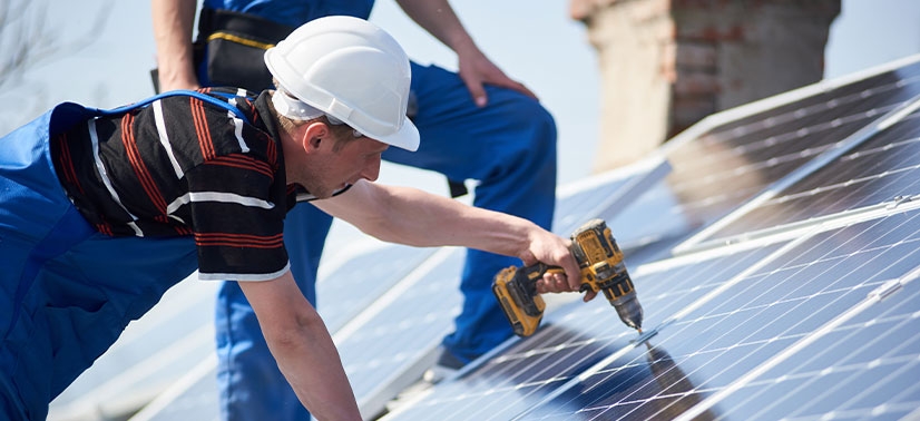 instaladores de painéis solares realizando o aperto de uma placa solar com uma parafusadeira em suas mãos enquanto outro é visto dando orientações