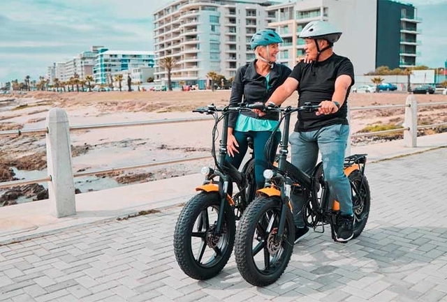 duas pessoas em cima de uma bicicleta elétrica e ao fundo a areia do mar e prédios
