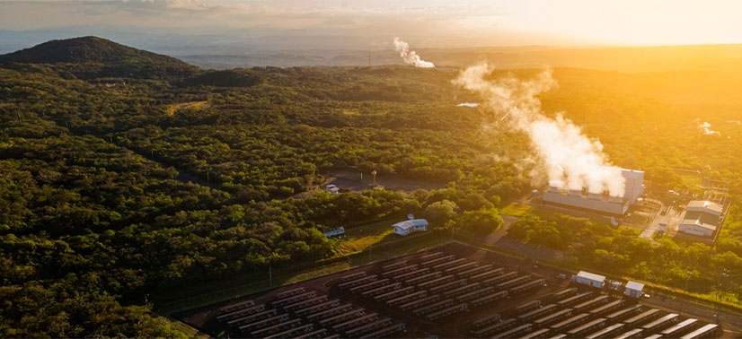 instalação industrial de energia geotérmica próxima a um campo de painéis solares, cercada por vegetação e montanhas