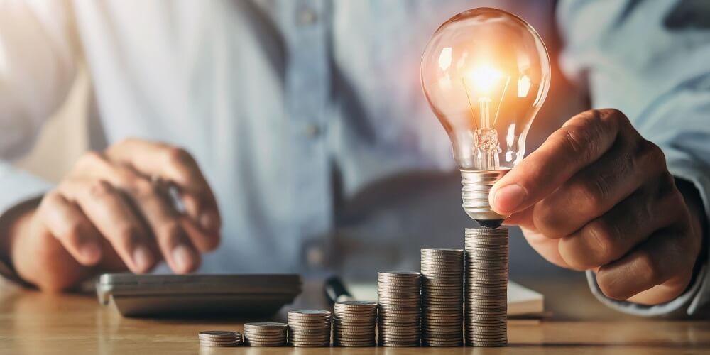 pessoa colocando uma lâmpada acesa sob uma pilha de moedas e ao fundo utilizando uma calculadora