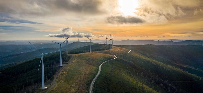 paisagem com um parque eólico situado sobre uma colina. Há várias turbinas eólicas alinhadas ao longo do caminho sinuoso no topo da colina.